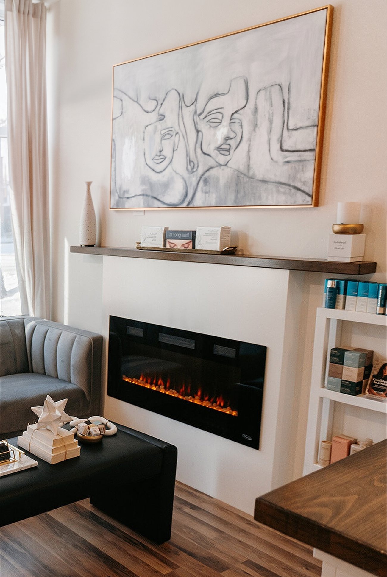 The lounge at Elegance Medispa in Bridgeland: a line-drawn painting of faces in a gold frame above a mantel and fireplace, grey velvet chairs, skincare on white shelves